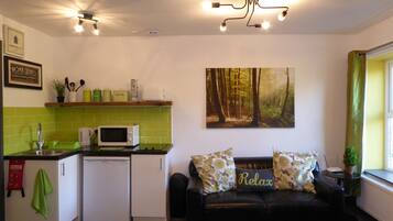 A compact kitchen with a microwave, a small refrigerator, and a sink. There's a green-tiled backsplash, a black leather sofa with 'Relax' pillows, and a forest-themed painting on the wall.