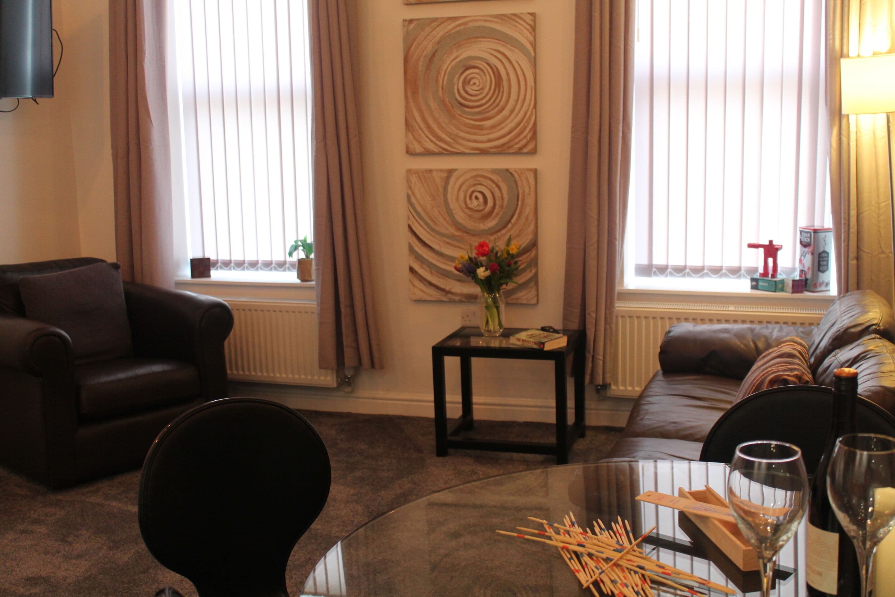 Un salon avec un canapé en cuir marron, une table basse en verre ornée de verres à vin et d’un vase de fleurs, et un fauteuil noir.