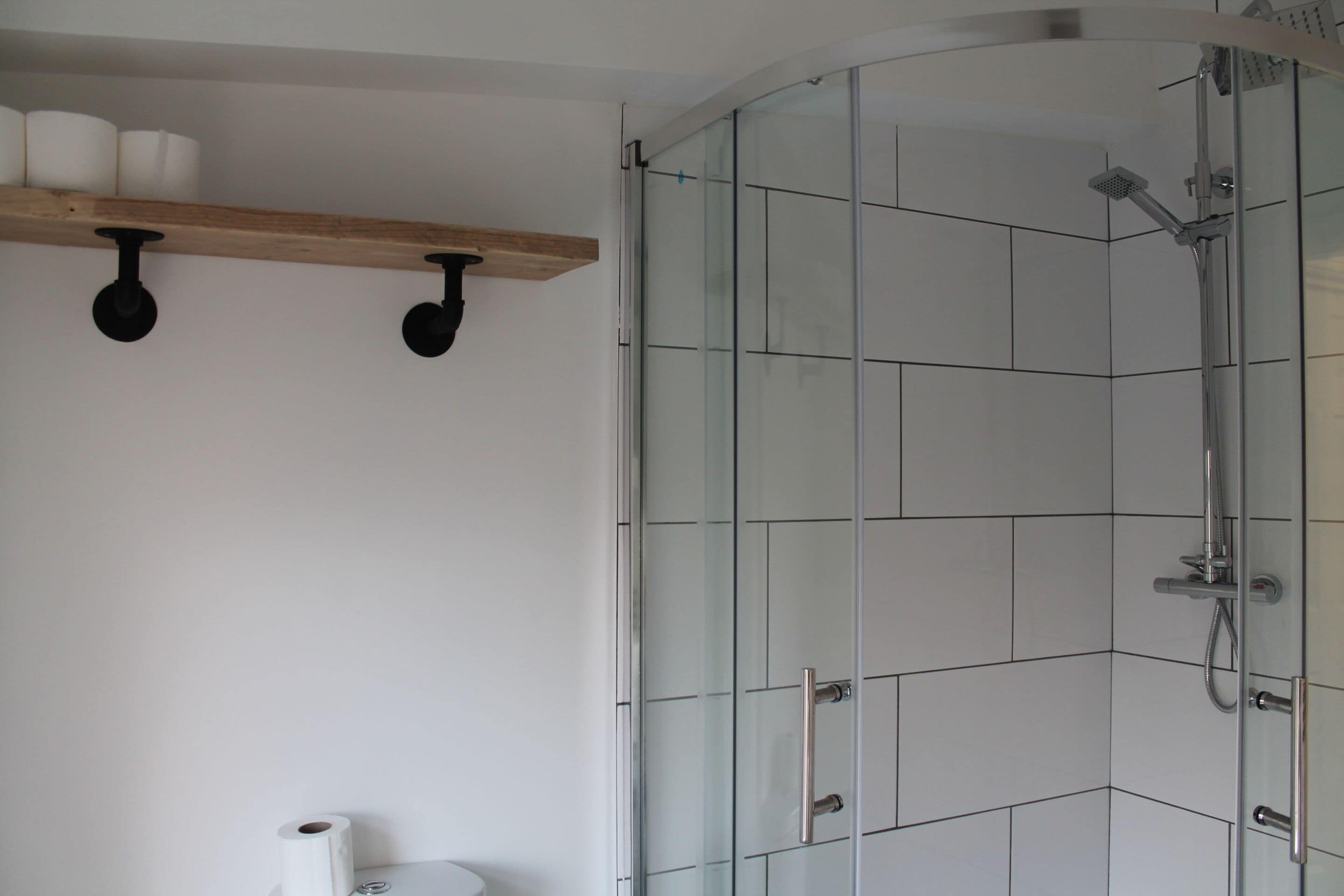 A bathroom with a glass shower enclosure, a wooden shelf with towels, and a toilet.