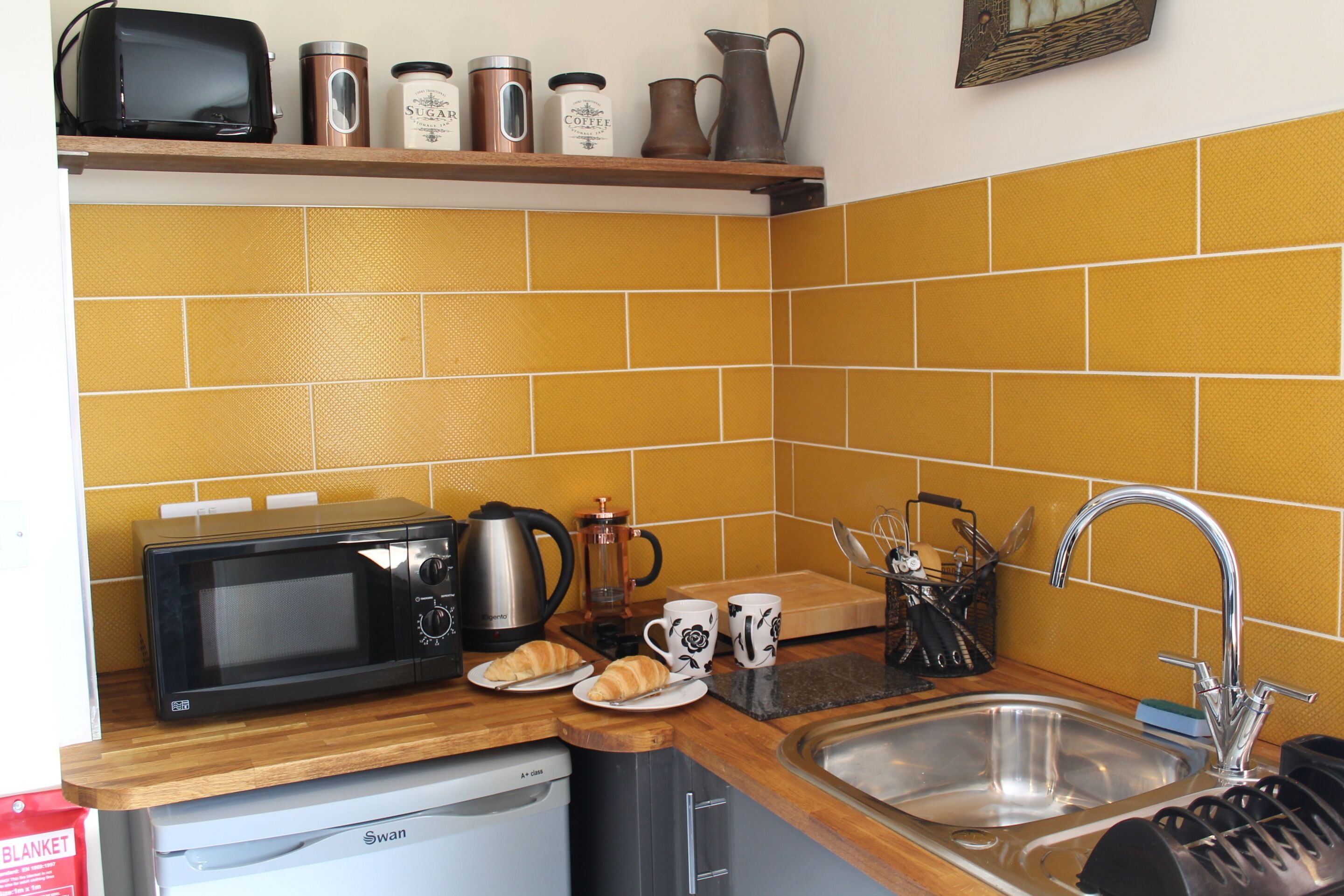A kitchen with a microwave, coffee maker, and toaster on the counter, a sink, and a shelf with jars and a pitcher.