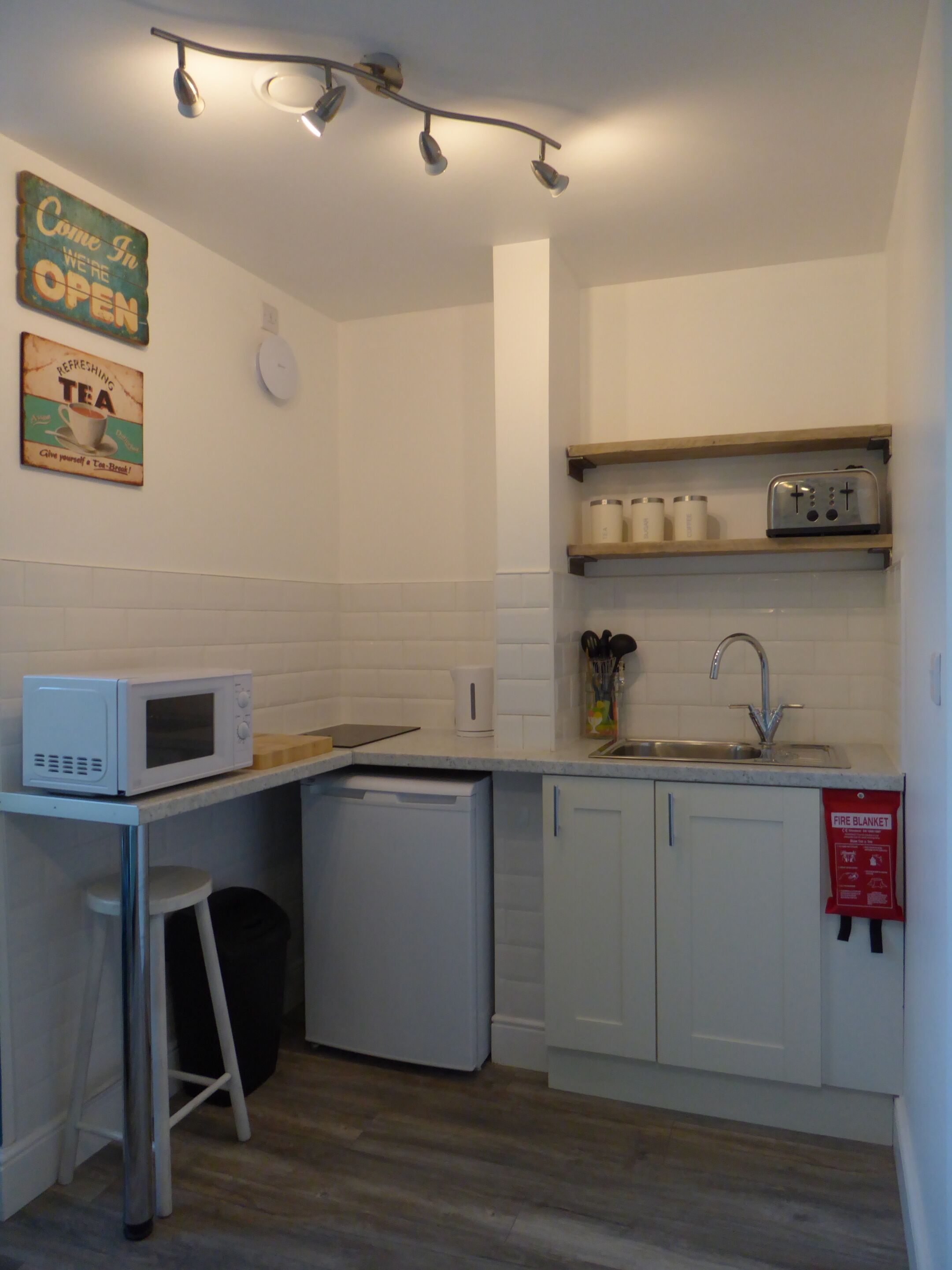 A compact kitchen with a microwave, toaster, and sink. There are two stools and a fire blanket on the wall.