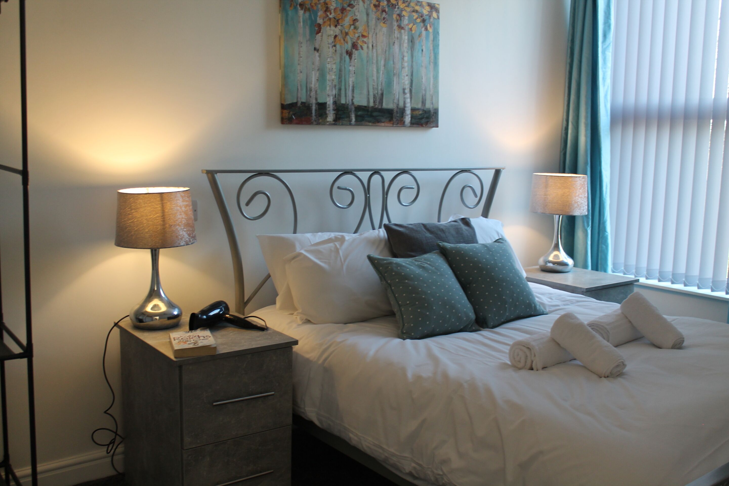 A neatly made bed with pillows and towels, a bedside lamp, a phone, and a small table with a lamp.