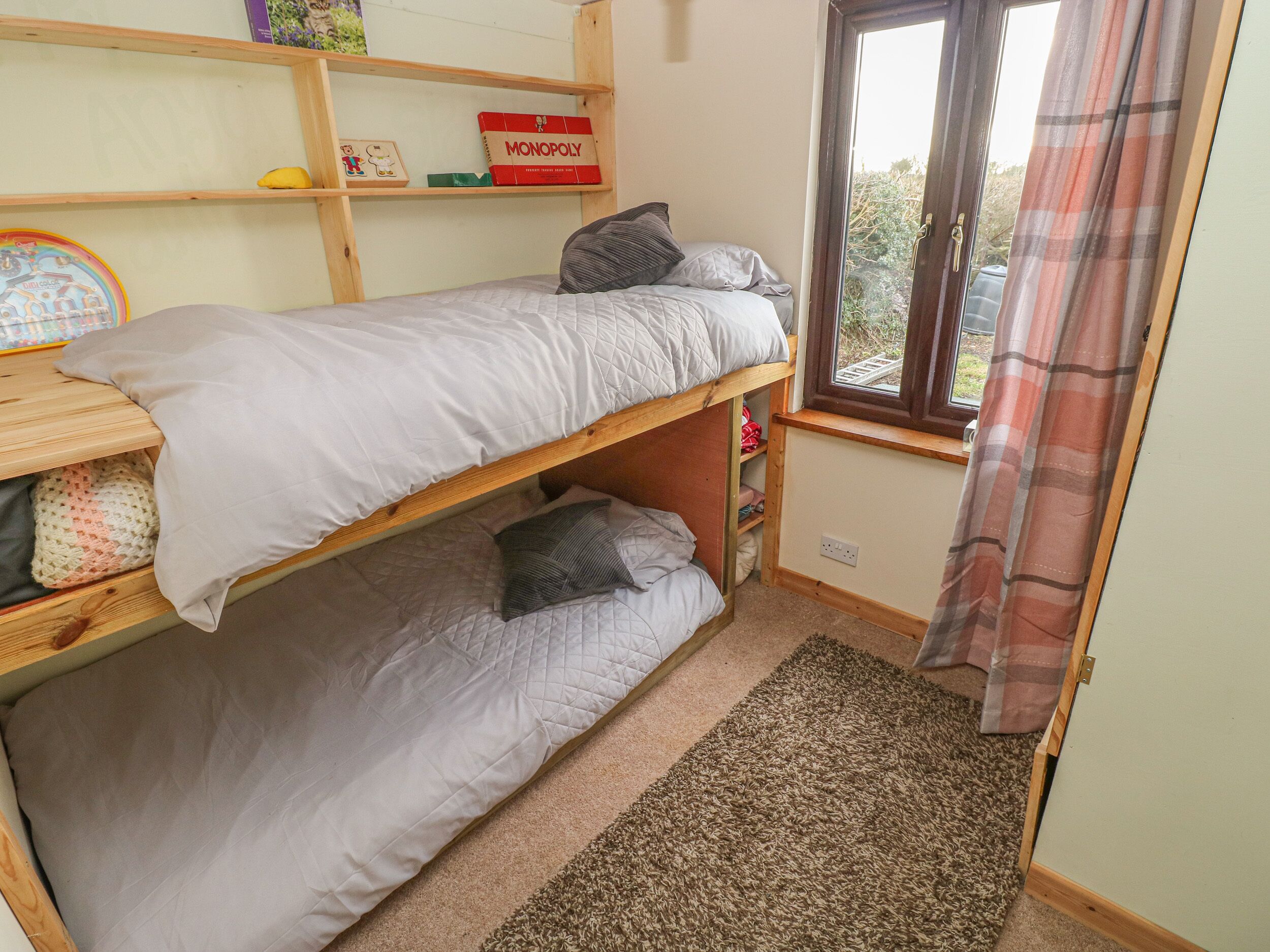 A dormitory room with two bunk beds, a Monopoly board game, a window with a view of greenery, and a plaid curtain.