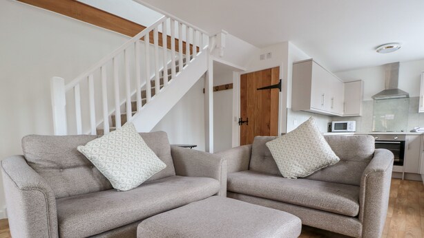 A modern living room with two grey sofas and an ottoman, a wooden staircase, and a kitchen area.