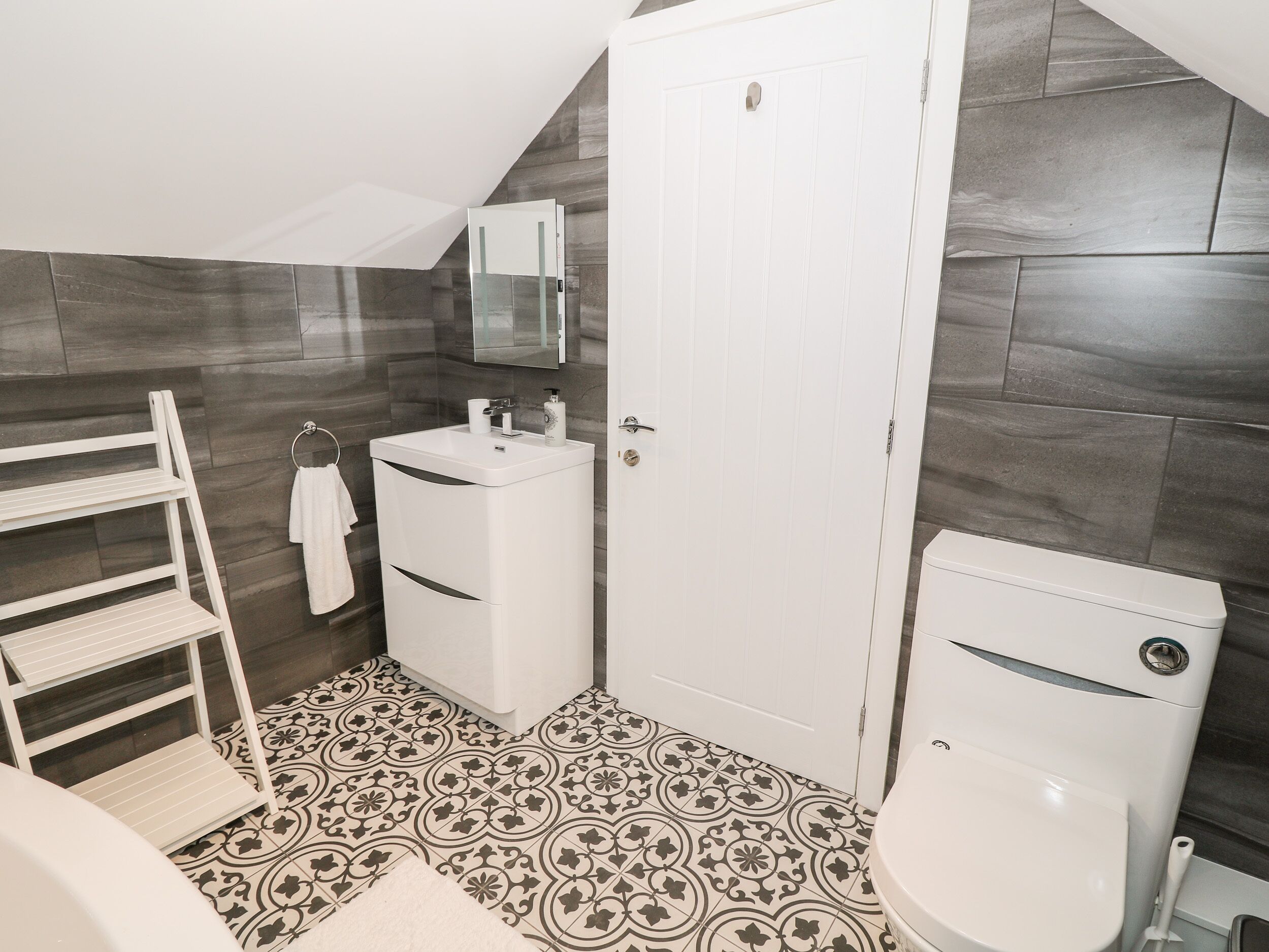 Een kleine, moderne badkamer met een witte wastafel, toilet en badkamermeubel. De vloer heeft een betegeld patroon, en er is een handdoekhouder met een witte handdoek.