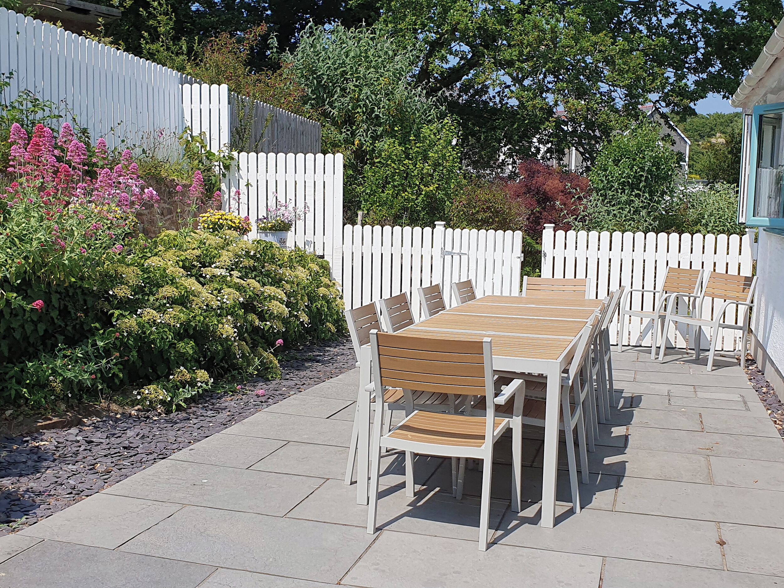 Une terrasse extérieure avec une table et des chaises en bois, entourée d’une clôture blanche en piquets et d’une végétation luxuriante.