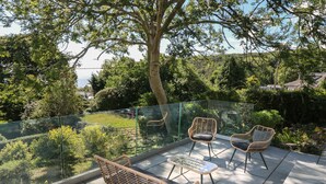 Een balkon met rieten meubilair, een glazen balustrade en uitzicht op een tuin en bomen.