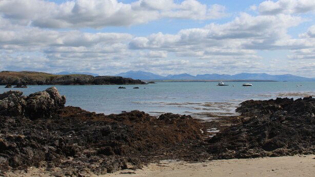 A coastal landscape with rocky shores, calm sea, and distant mountains under a partly cloudy sky. - The Granary - Rhoscolyn (Holyhead)
