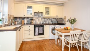 A compact kitchen with white cabinets, a microwave, a washing machine, and a dining area with two chairs and a table. - The Granary - Rhoscolyn (Holyhead)