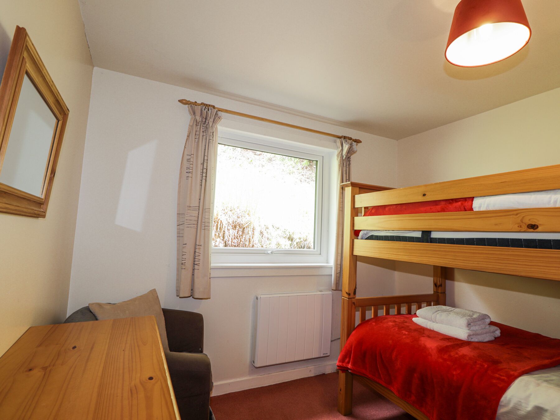 Une petite pièce bien rangée, avec un lit superposé, une table, une chaise, une fenêtre avec des rideaux et un radiateur.