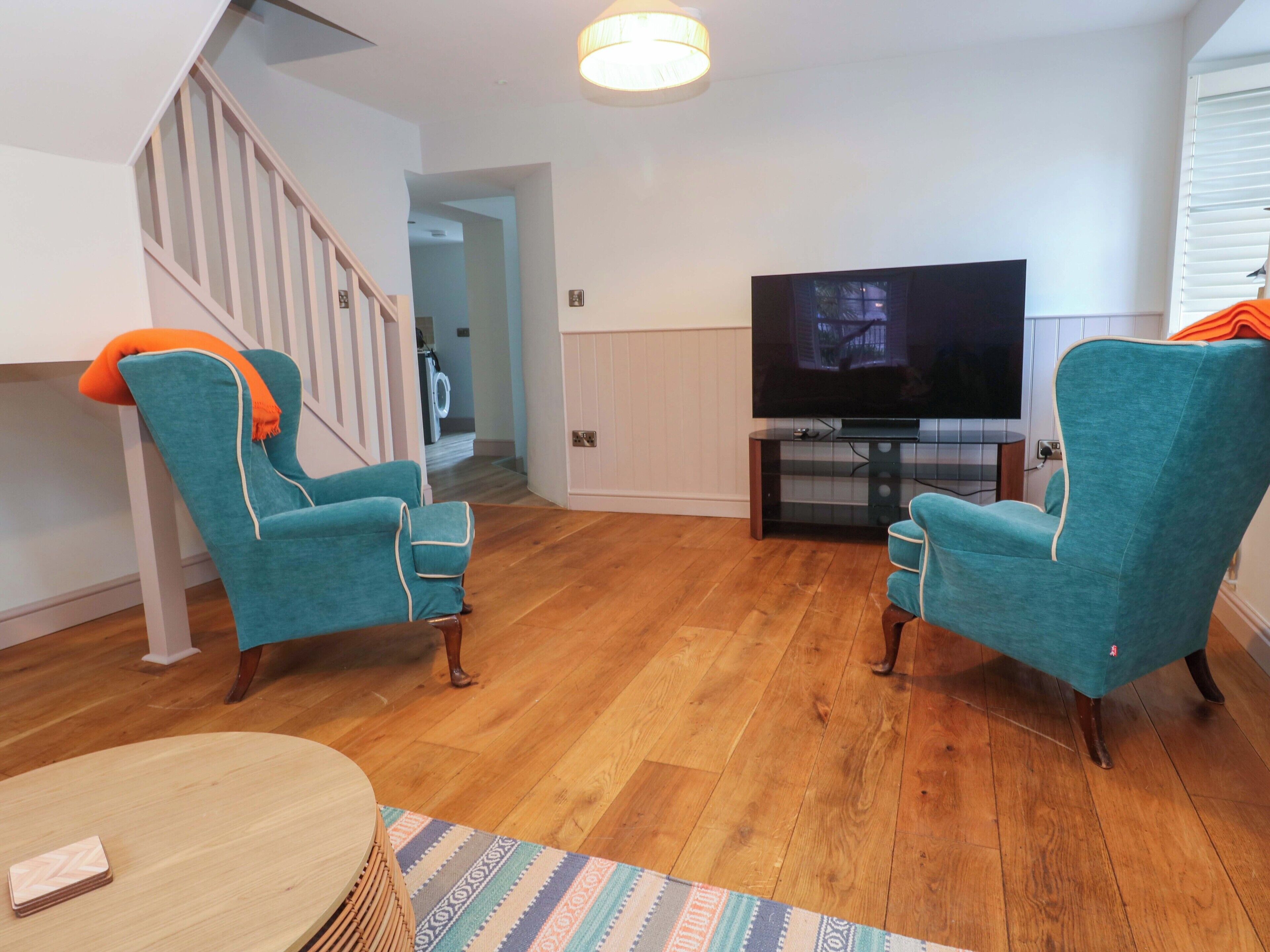 Una sala de estar con sillones azul verdoso, suelo de madera, un televisor sobre un mueble bajo y una escalera.