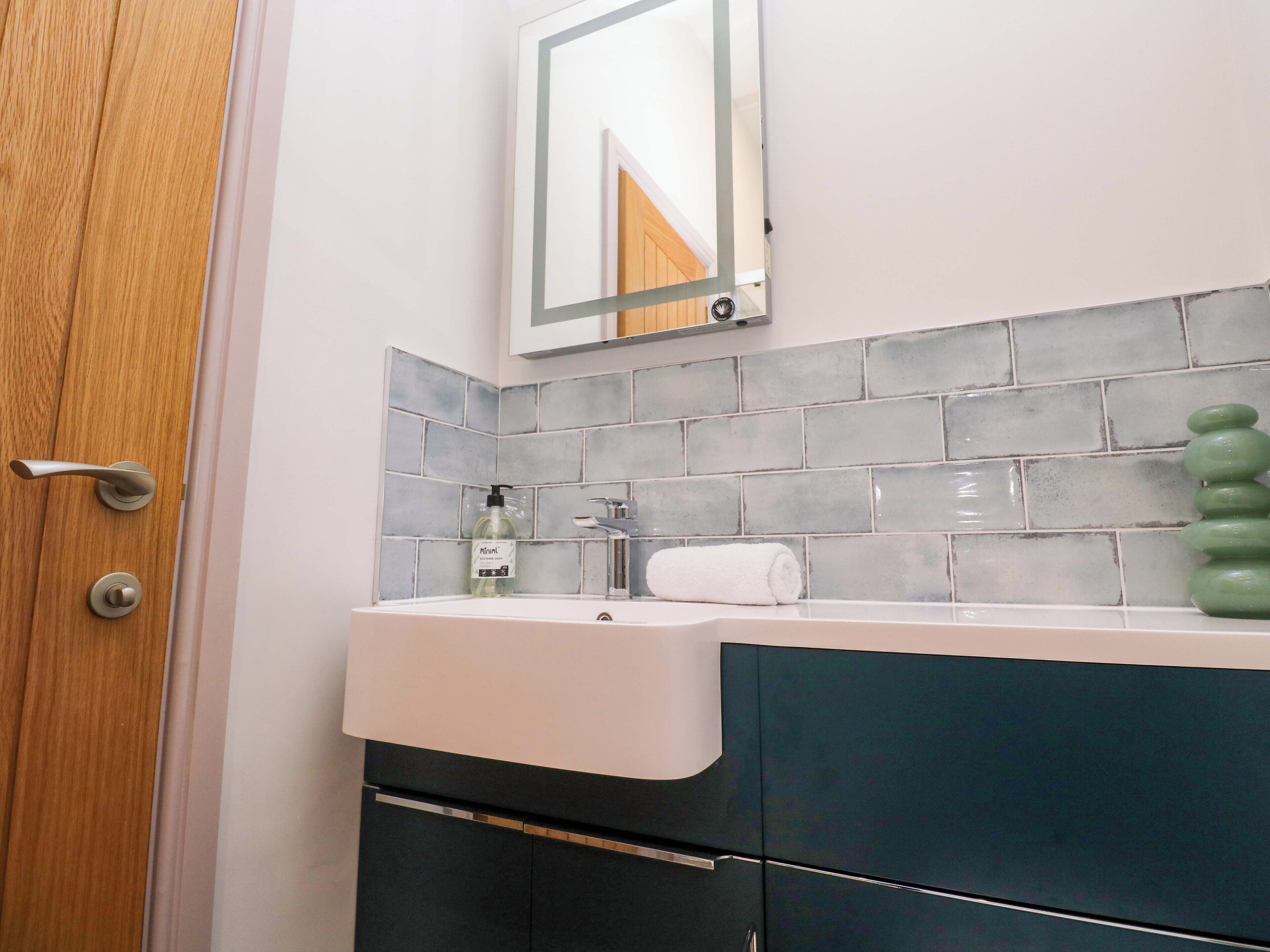Een badkamer met een houten deur, een witte wastafel, een spiegel en grijs betegelde wanden.