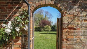 A brick archway with a metal pipe, leading to a grassy lawn with trees and a hedge. - Bakehouse Farm (Princes Risborough)