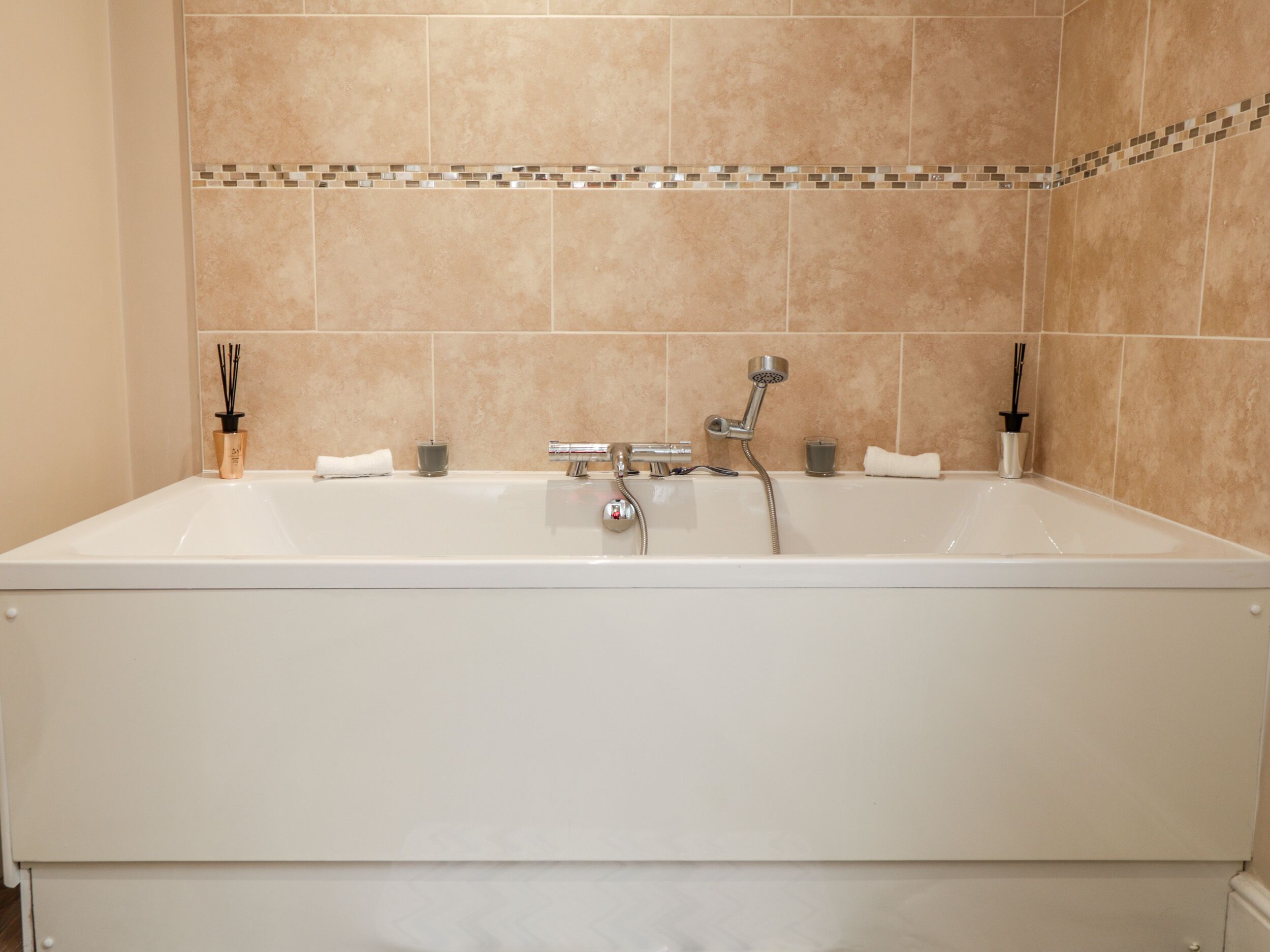 Eine große, rechteckige Badewanne mit chromfarbenen Wasserhahn und Duschkopf, vor beige gefliesten Wänden mit dekorativer Bordüre.