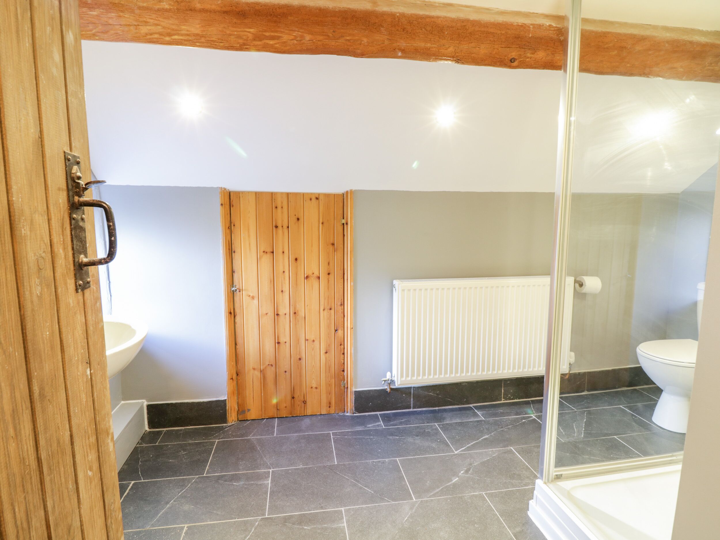 Une salle de bain avec une porte en bois, un radiateur blanc, une cabine de douche en verre et un sol carrelé de carreaux noirs.