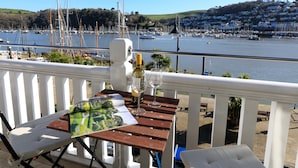 A balcony with a table set for two, overlooking a marina with boats and a hilly town in the background. - 2 Embankment House (Dartmouth)