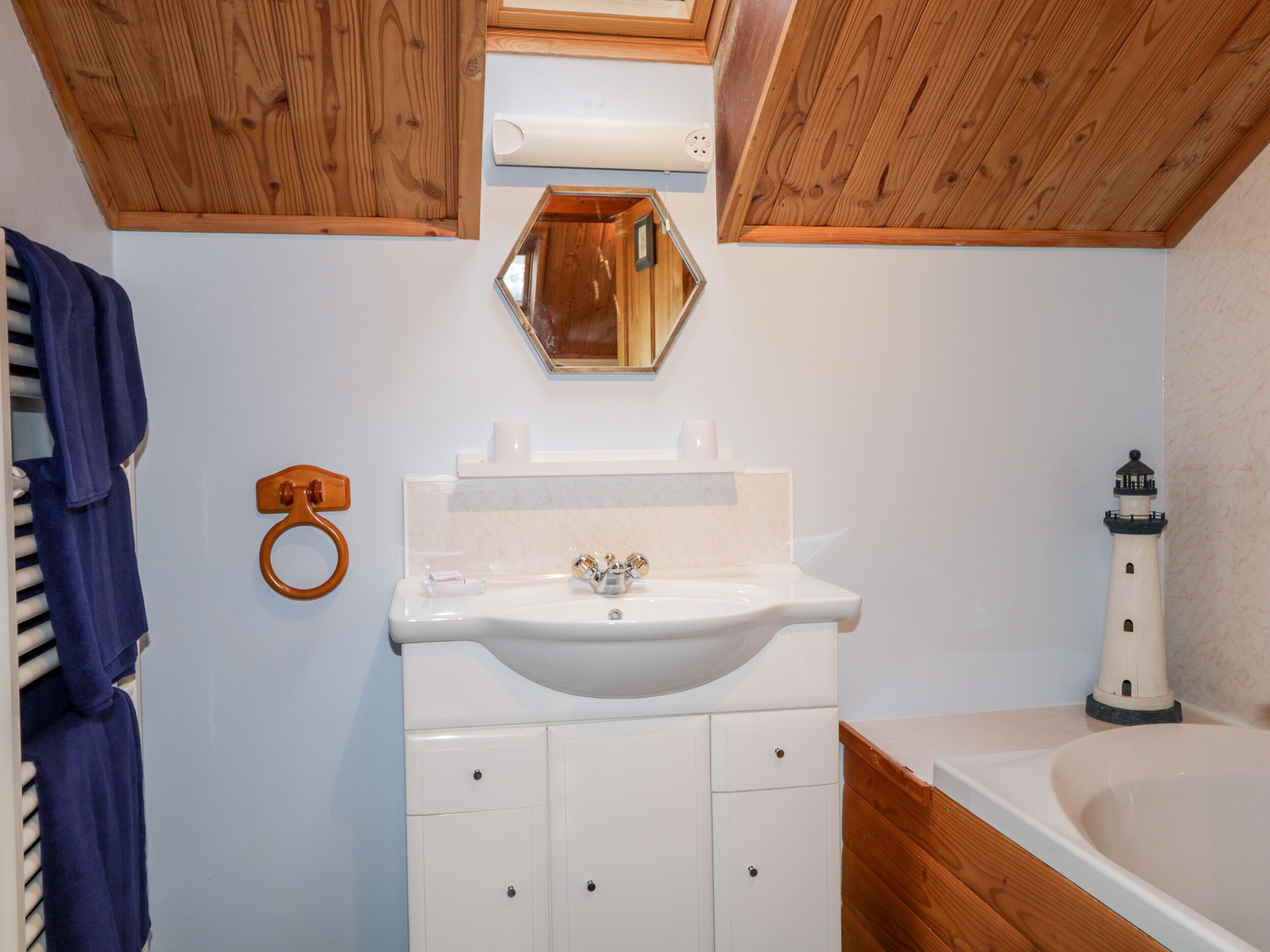Une salle de bain avec un plafond en bois, un lavabo blanc avec un miroir au-dessus, un porte-serviettes et une maquette de phare.