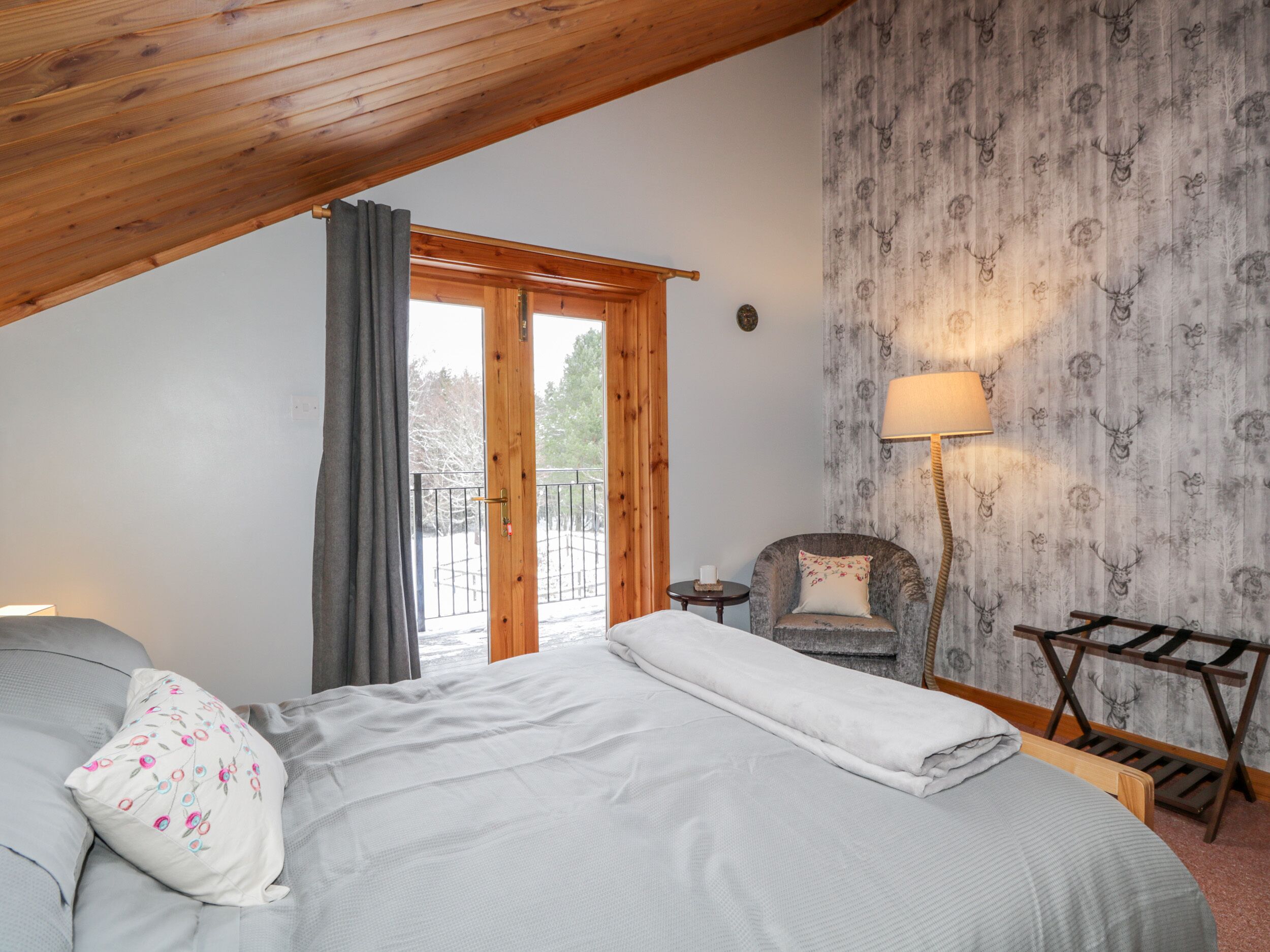 Ein Schlafzimmer mit Holzbalkendecke, einem Bett mit weißer Decke, einem Sessel mit Blumenkissen, einem kleinen Tisch und einer Lampe.