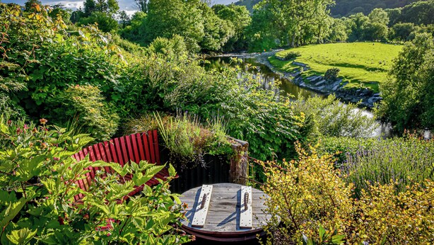 En flodsidehave med en træterrasse, frodigt grønt og udsigt til en bakke i baggrunden.