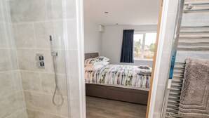 A modern bathroom with a glass-enclosed shower, a wooden floor, and a visible bed with a colorful bedspread in the adjacent room. - Beechnut Cottage (Tewkesbury)
