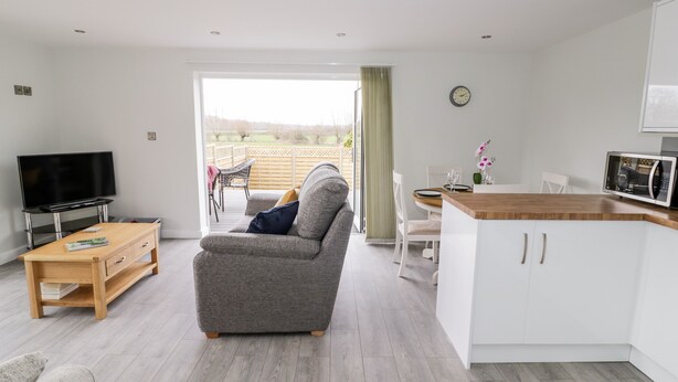A modern living room with a grey sofa, wooden coffee table, and a flat-screen TV. A kitchen area with white cabinets and a microwave is visible. - Beechnut Cottage (Tewkesbury)