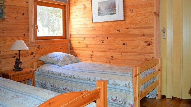 A cabin-style room with two wooden bunk beds, a window, a framed picture on the wall, and a lamp on a nightstand.