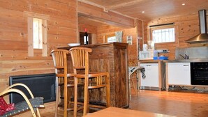 A wooden cabin interior with a kitchen area, a dining table, and two wooden chairs.