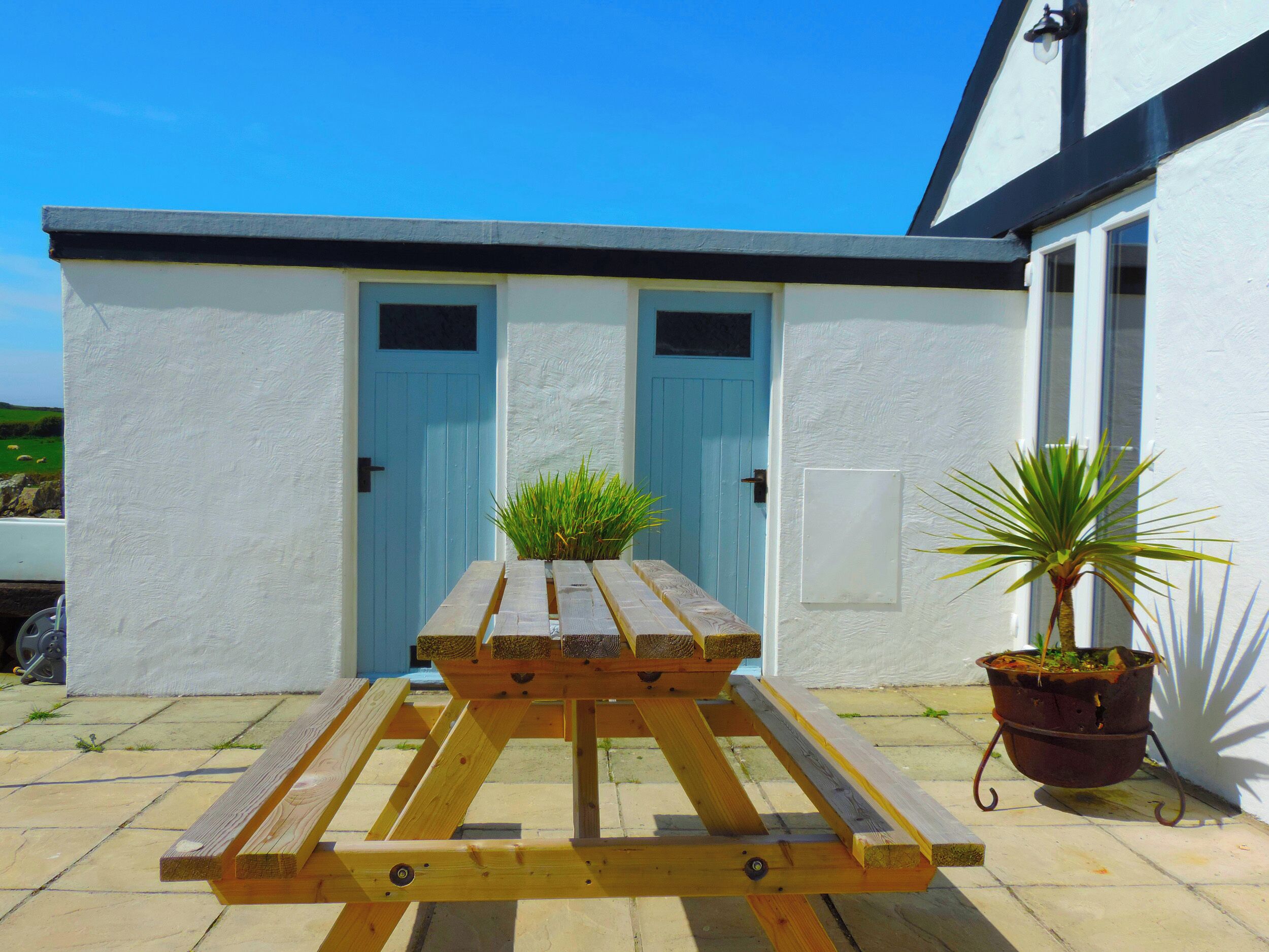 Une table de pique-nique en bois, avec un pot de fleurs à côté d’un bâtiment blanc aux portes bleues.