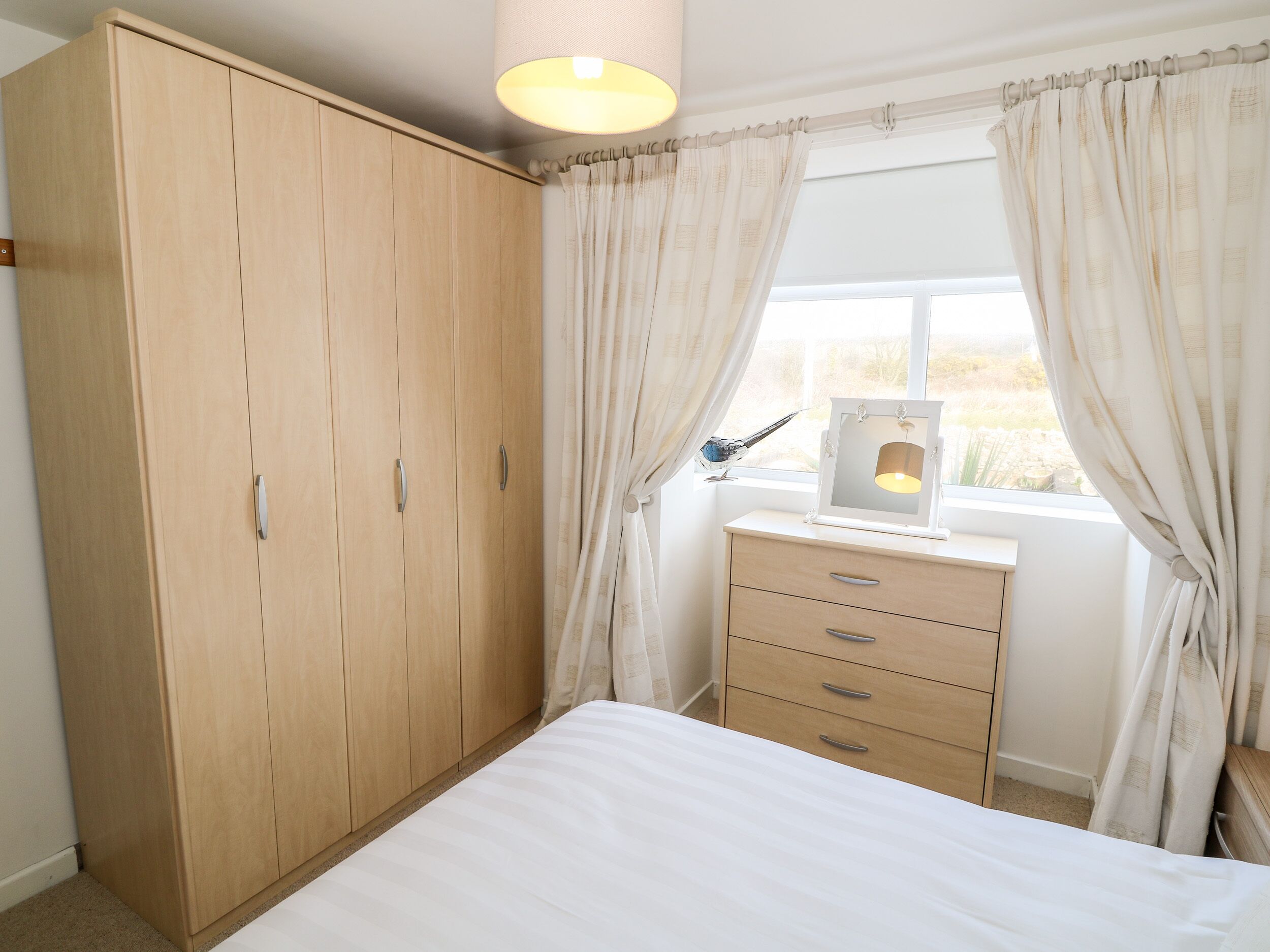 Une chambre avec une armoire en bois, une table de chevet avec une lampe et une fenêtre avec des rideaux.