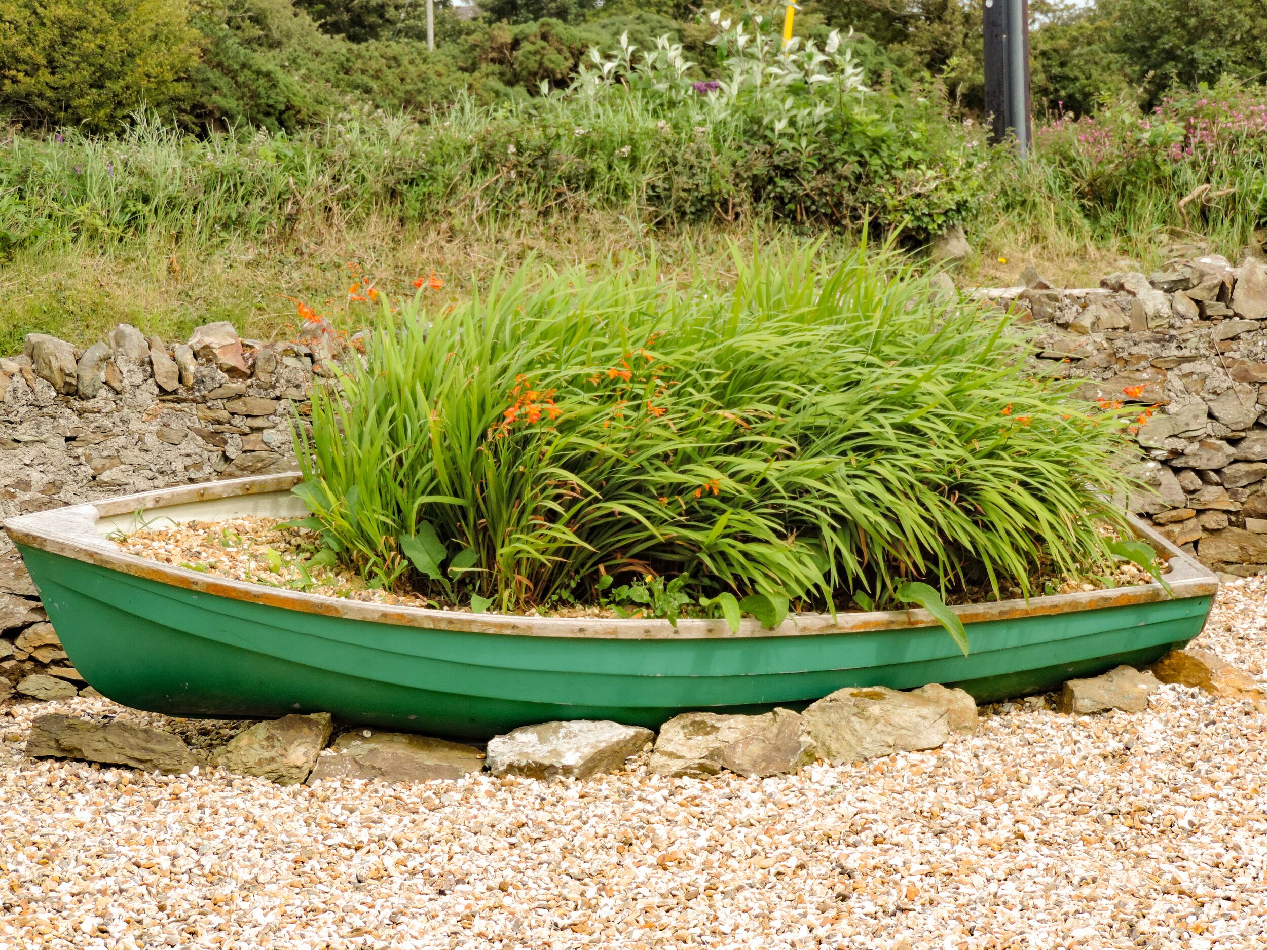 A green boat-shaped planter filled with various plants and flowers, surrounded by a gravel path and a stone wall.