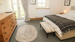 Une chambre à coucher comprenant un lit, une commode en bois, un tapis rond en fibres tressées, un banc, une fenêtre avec des rideaux et un radiateur.