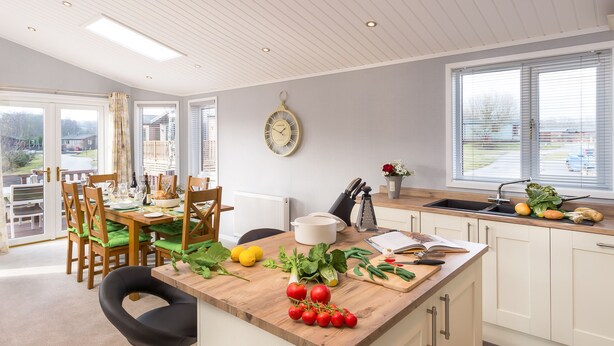 Une cuisine moderne avec un îlot central en bois, un coin repas avec des chaises et une table, et une vue sur l’extérieur grâce à de grandes fenêtres.