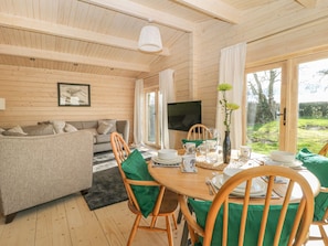 A cozy room with a wooden ceiling, a sofa, a dining table with chairs, a TV, and a window with a view of greenery.