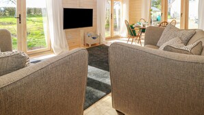 A cozy living room with a flat-screen TV, two sofas, a dining table with chairs, and large windows offering a view of the outdoors.