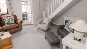 A living room with a brown sofa, a grey armchair, a grey ottoman, and a white table with a lamp. There is a staircase with white railings and a door with frosted glass panels. - Kite Cottage (Brackley)