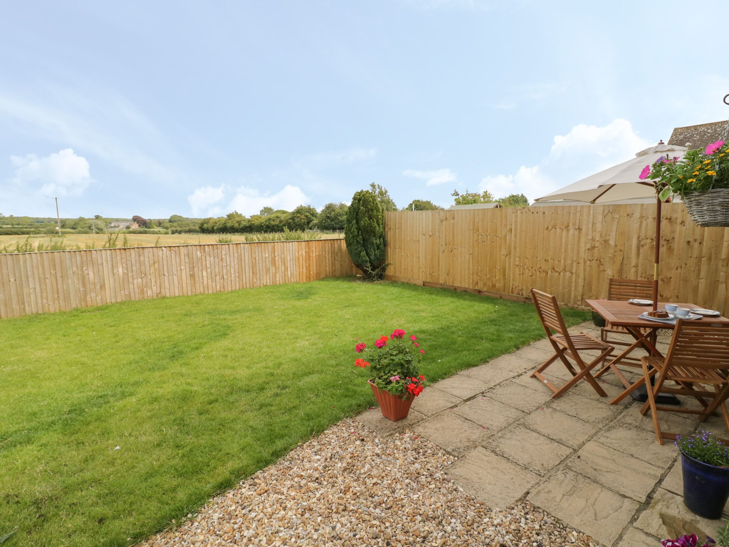 A backyard with a wooden fence, a lawn, a patio area with a table and chairs, and a potted plant with flowers.