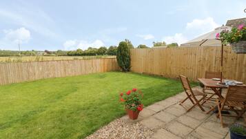A backyard with a wooden fence, a lawn, a patio area with a table and chairs, and a potted plant with flowers.