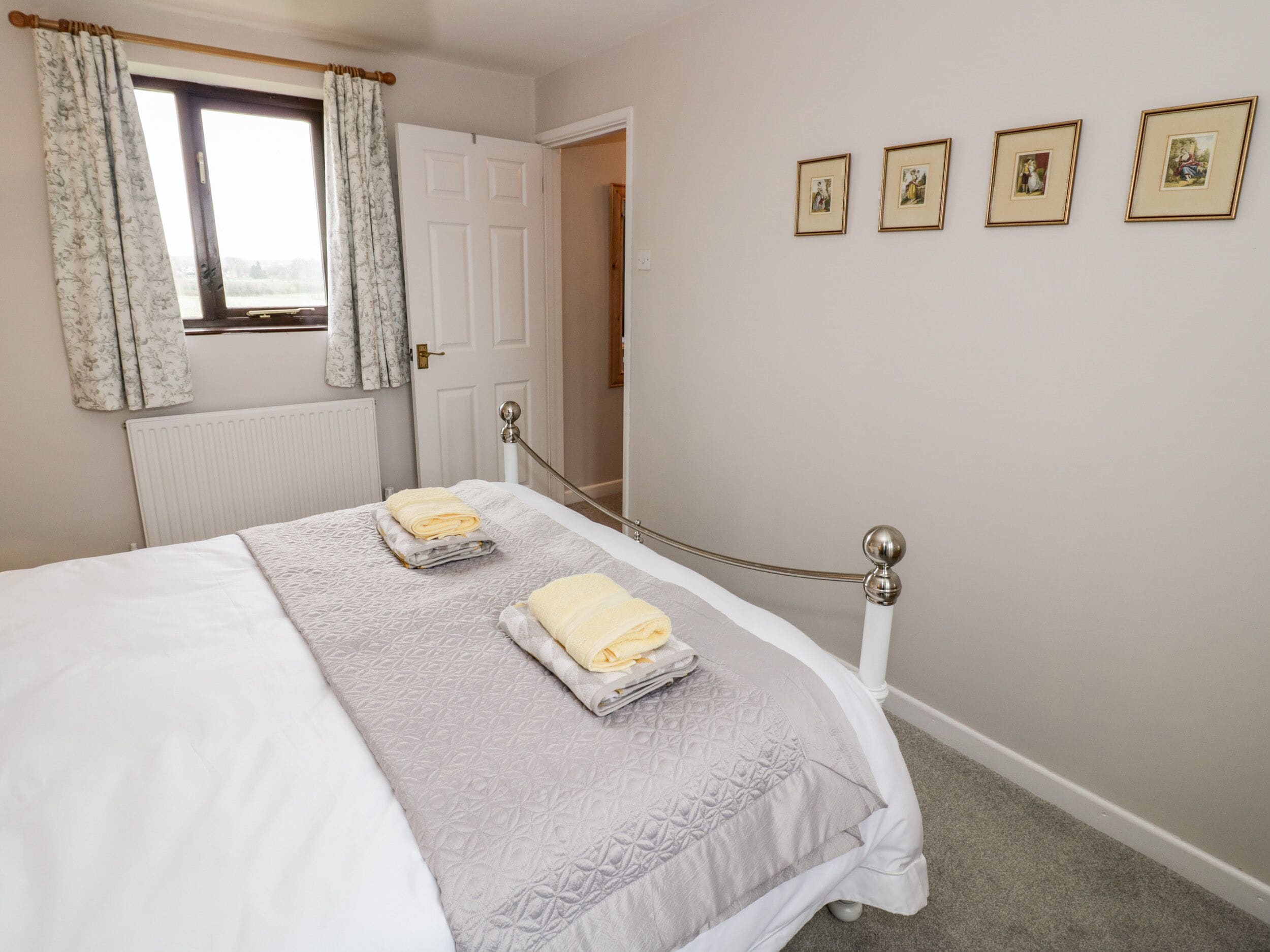 A neatly made bed with a grey quilt and two folded towels, a window with floral curtains, and a door with a view of the outside.