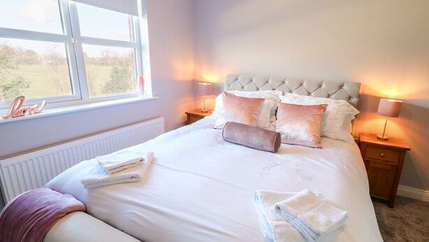 Een net opgemaakt bed met kussens en een opgerolde handdoek, een nachtkastje met een lamp, en een raam met uitzicht op het groen.