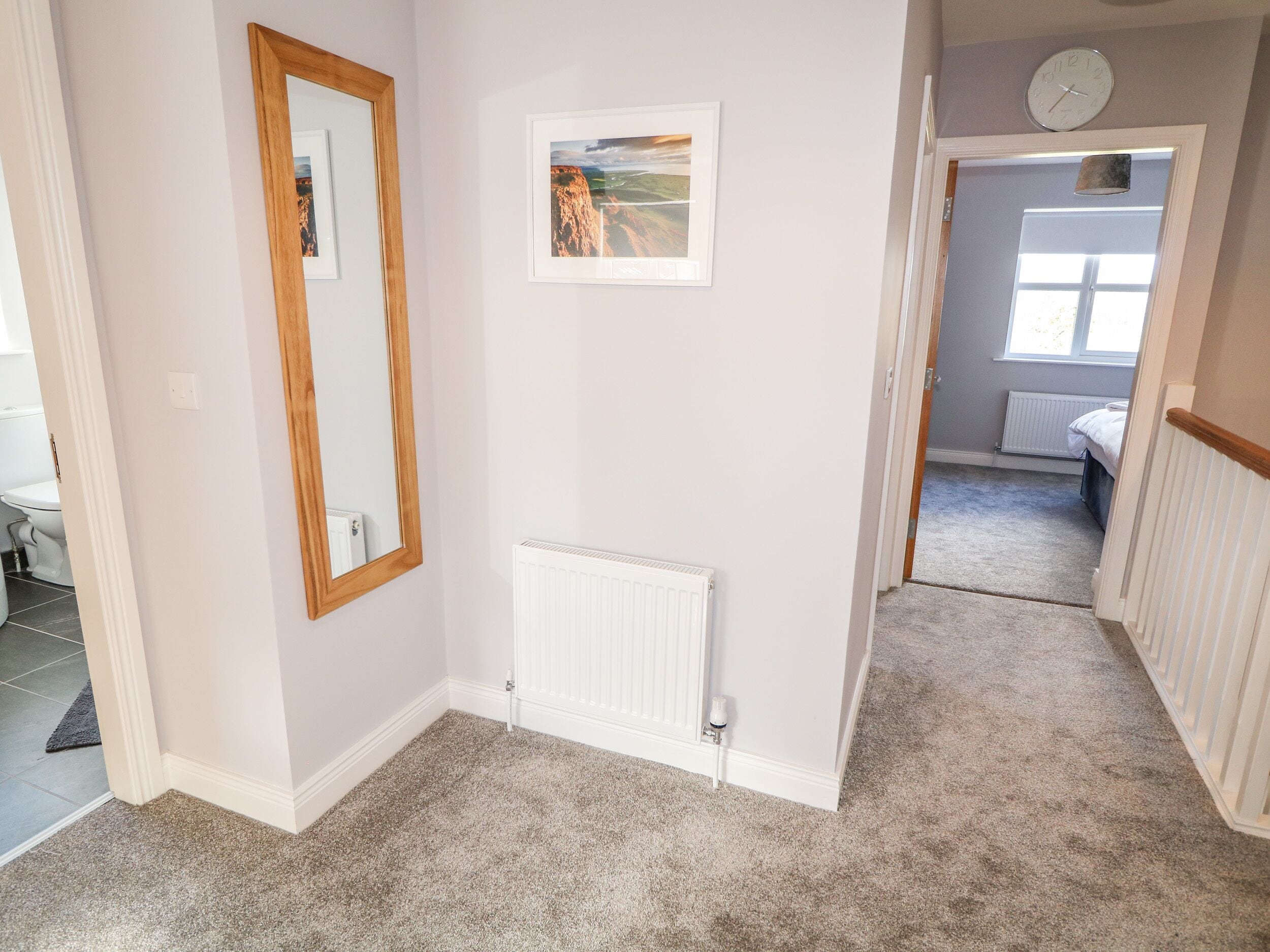 A hallway with a wooden-framed mirror, a radiator, and a framed landscape painting on the wall.