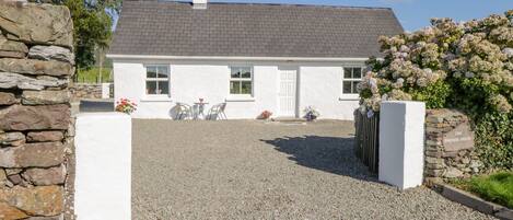 A white house with a gravel driveway, stone walls, and a garden with flowers.