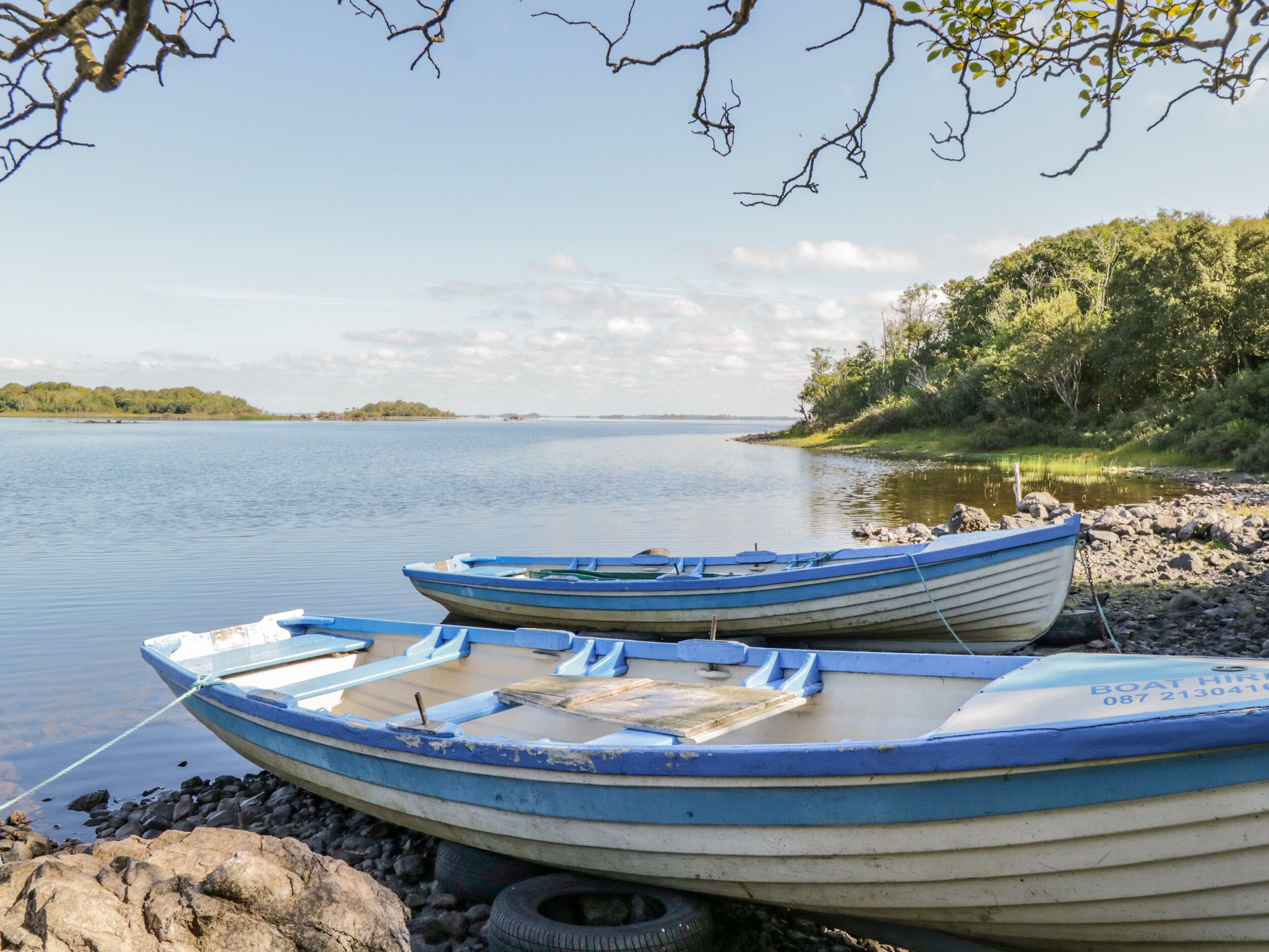Two boats tied to a rocky shore by a calm lake with a forested island in the background.
