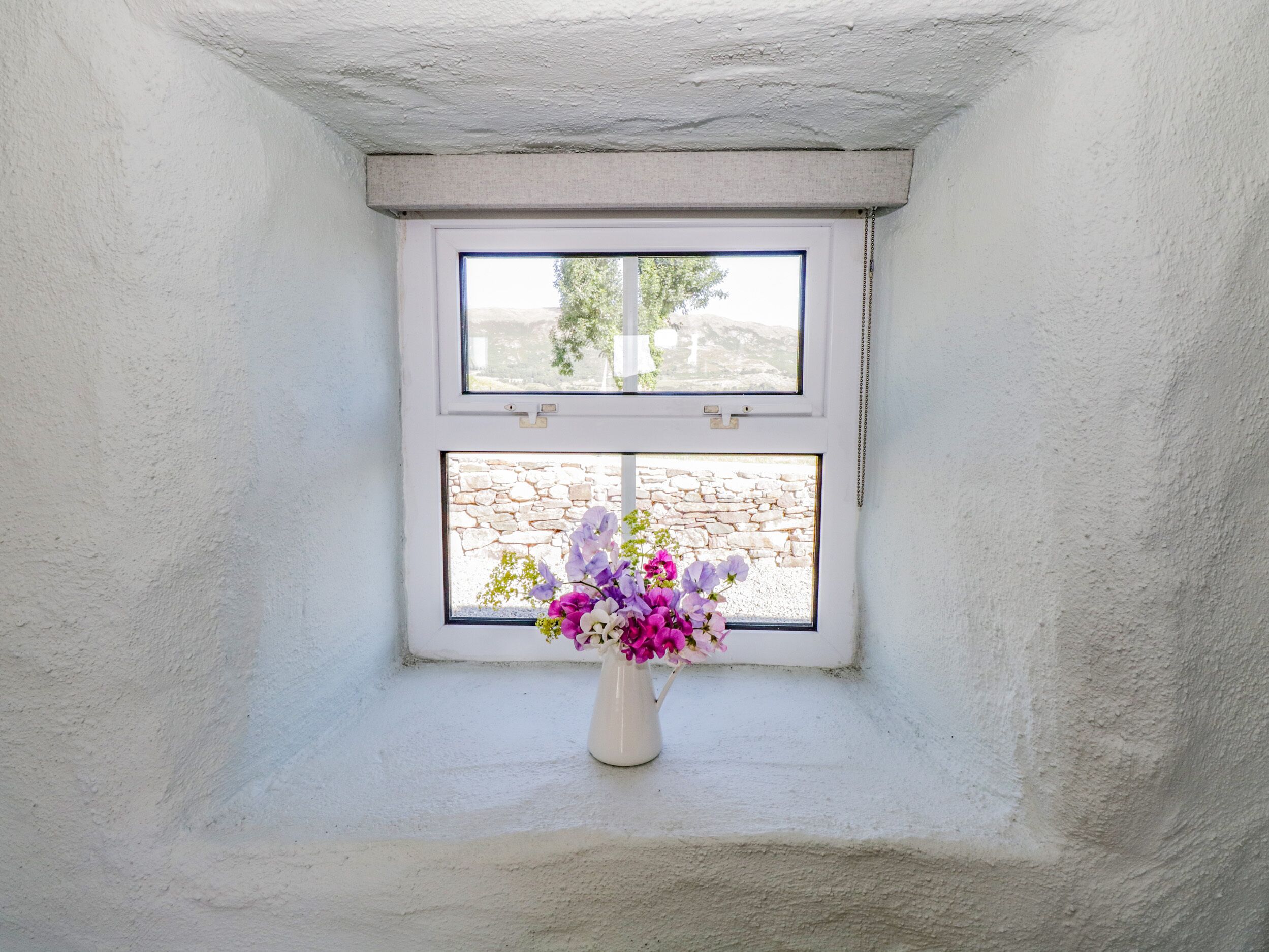 Een wit raam met uitzicht op een stenen muur, een witte vaas met bloemen en een wit plafond.