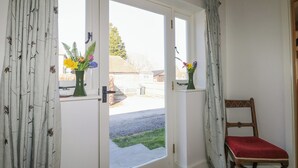 Habitació amb una porta corredissa de vidre que dona a un jardí, una cadira de fusta amb coixí vermell i dos vasetes amb flors al llindar de la porta.