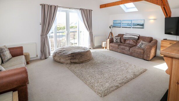 A spacious living room with a high ceiling, wooden beams, a large window, a brown leather sofa, a beige rug, and a television. - Bay View (Holyhead)