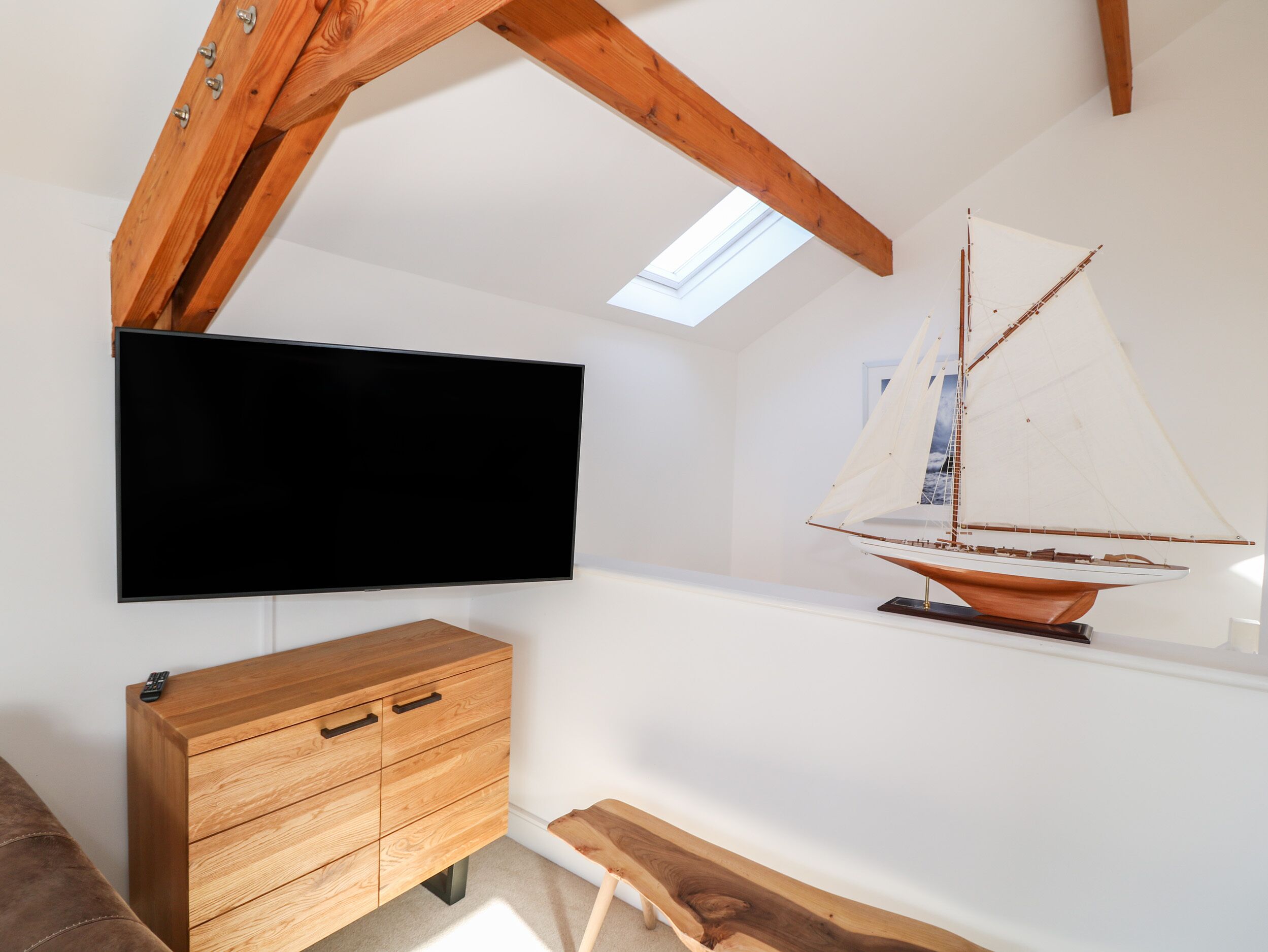 Ein Zimmer mit an der weißen Wand montiertem Fernseher, einem Holzschrank mit Schubladen, einem Modellsegelboot und einer geneigten Decke mit Dachfenster.