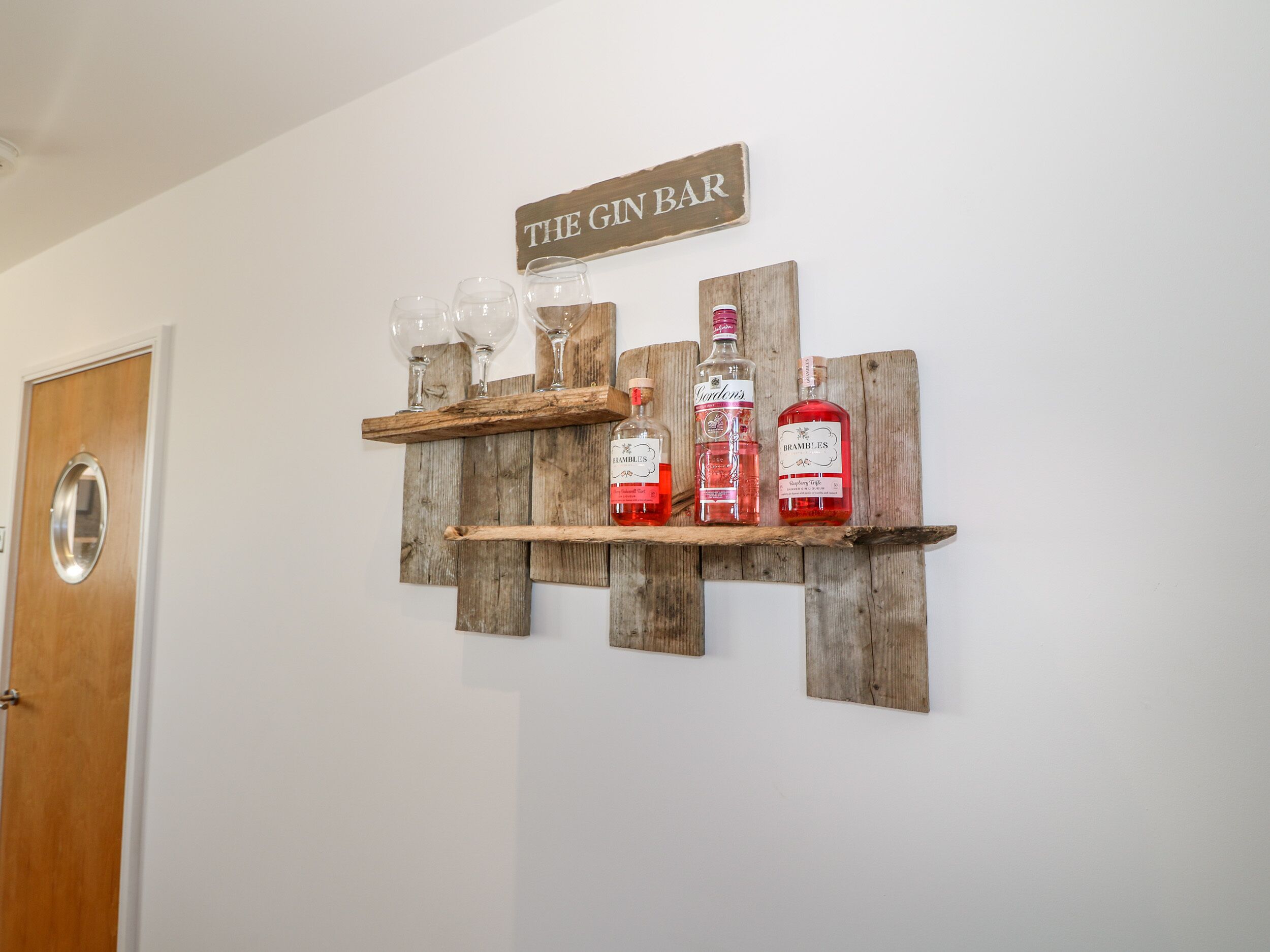 A wooden wall-mounted shelf with three bottles labeled 'Brambles' and 'Gordon's,' glasses, and a sign reading 'THE GIN BAR.'