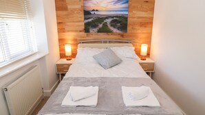A neatly made bed with two white towels folded on it, a grey pillow, and a wooden headboard with a picture of a beach scene. - Bay View (Holyhead)