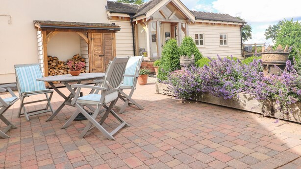 Un patio al aire libre con mesa y sillas, una pérgola de madera y un jardín con flores moradas.