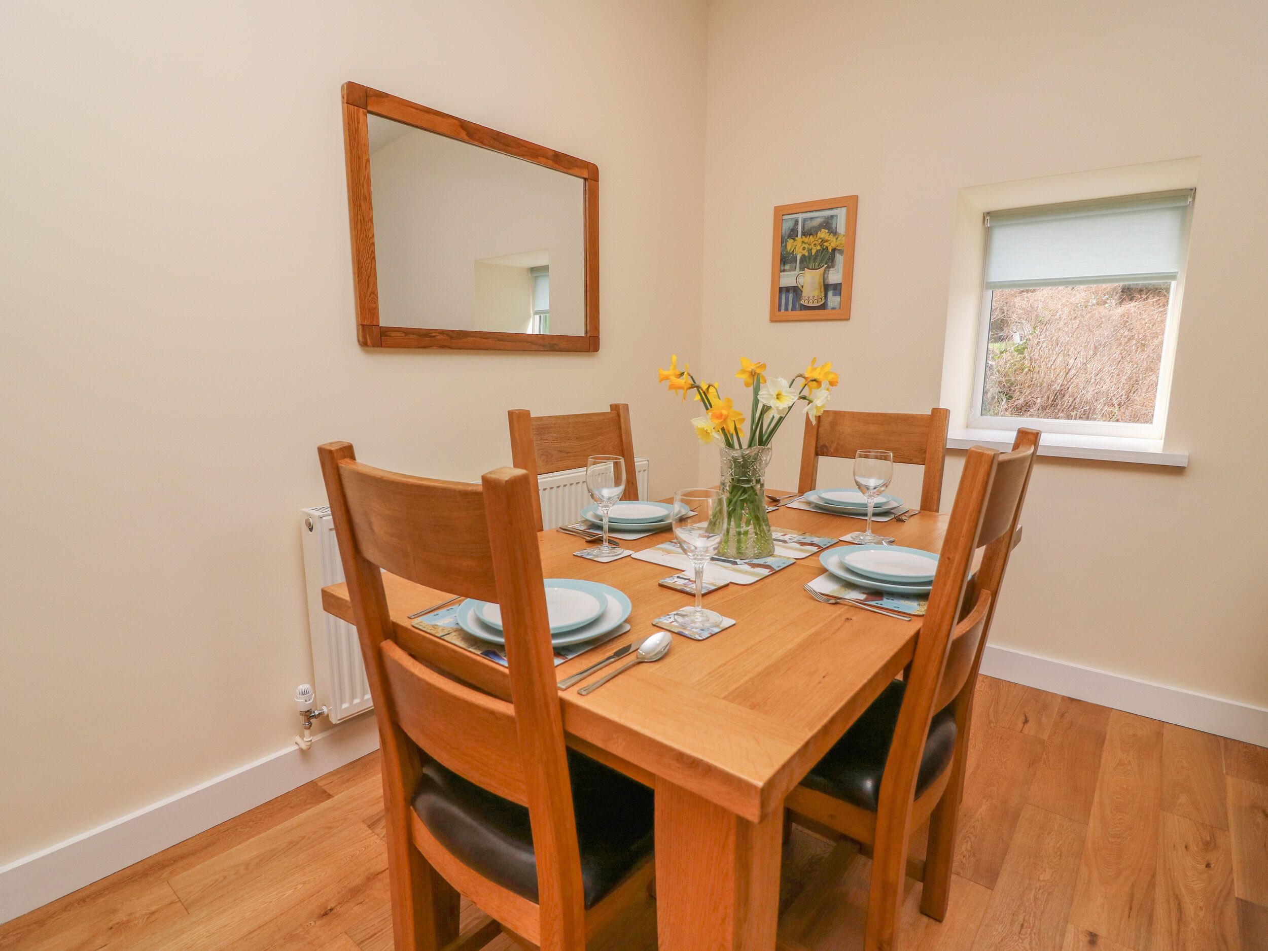 A dining room with a wooden table set for four, chairs, a vase with flowers, a painting, and a window with a view.